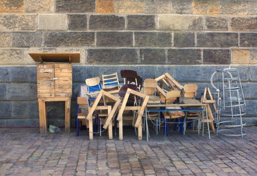 A collection of wooden chairs of various styles and finishes, including natural, painted, and stained, are stacked and arranged against a weathered brick wall outdoors. The chairs include both traditional and modern designs, some with armrests and others with simple backrests. To the left, there is a small wooden cabinet with a hinged lid and a slatted front, appearing aged and slightly worn, positioned on the cobbled pavement surface. On the right, a metal shelving unit with a curved wireframe structure is partially visible. The scene appears to be part of a garden clearance or salvage operation, situated on a paved outdoor area, possibly in Wimbledon, with natural daylight illuminating the objects and casting soft shadows. The arrangement and condition of the furniture suggest they are awaiting disposal, re-use, or recycling as part of professional rubbish removal services handled by Garden Clearance Wimbledon, serving local properties in the SW19 postal district.