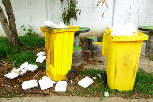 Two bright yellow wheeled rubbish bins made of plastic with textured surfaces are positioned on a pavement next to a white wooden fence, with one bin slightly in front of the other. The bins are open at the top, and scattered white disposable food containers with raised lids, some crumpled or knocked over, are spilling onto the ground around them. The surrounding environment includes patches of grass and soil, with a tree trunk visible on the left and a small patch of greenery climbing the fence in the background. The scene appears to take place outdoors in a residential or garden area, with natural daylight illuminating the scene. This image relates to rubbish removal and waste management, as represented by the scattered containers and the presence of waste bins, which may be serviced or emptied by a local waste disposal company such as Garden Clearance Wimbledon operating in the Wimbledon or SW19 postcode area.