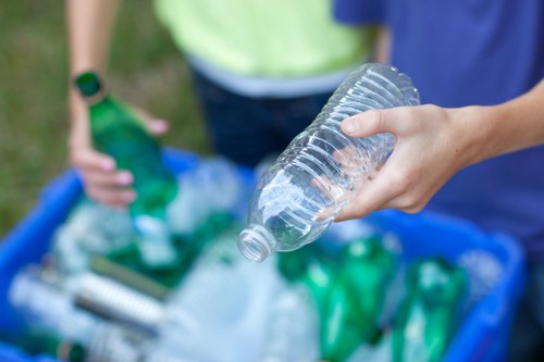 A close-up of a person's hand holding a clear plastic bottle, with a focus on the bottle's ridged body and open neck. In the blurred background, another individual is seen holding a green plastic bottle, near a blue recycling bin filled with various discarded plastic bottles and containers. The scene takes place outdoors on a grassy area, possibly in a garden or yard, suggesting a rubbish collection or recycling activity consistent with waste management services offered by Garden Clearance Wimbledon. The lighting is natural, highlighting the transparency of the plastic bottles and the textures of the items in the bin, emphasizing the focus on waste separation and disposal in a typical domestic or small-scale commercial context within Wimbledon or nearby areas.