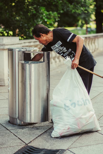 A person wearing a green glove on each hand is holding open a large, black, plastic rubbish bag that is partially filled with waste. The person is dressed in a casual, checked shirt with shades of yel