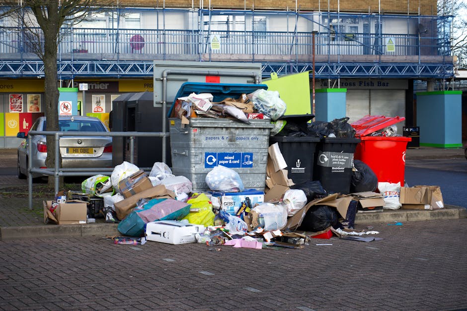 A black wheeled rubbish bin labeled 'ST. JOHN'S' positioned on a residential pavement at night, with its lid open and overflowing with various waste items including cardboard, plastic containers, and 