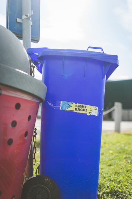 A row of multiple waste bins lined up outdoors beside a brick wall, with the closest bin in green featuring a textured lid and a white recycling symbol. Behind it, there are red and blue bins, all wit